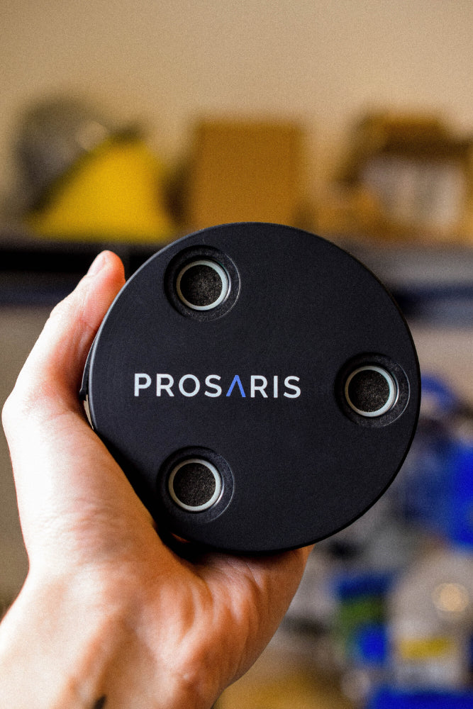 A hand is holding a PROSARIS OL1 device. A blurred industrial setting can be seen in the background.