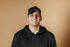 A smiling man in a black hat and hoodie in front of a light brown backdrop.