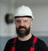 A bearded man wearing a white hard hat, protective eye-ware, and a black shirt.